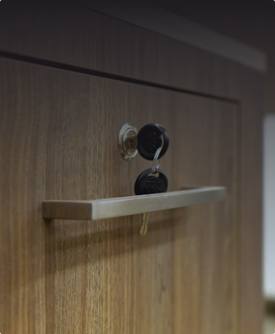 Zbliżenie na drzwi drewnianej szafki z metalową klamką i kluczem włożonym do zamka, z czarnymi plastikowymi osłonami kluczy.
