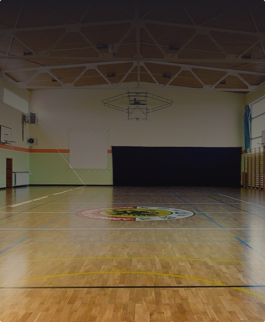 Pusta sala gimnastyczna z drewnianą podłogą, oznaczeniami sportowymi, koszem do koszykówki i dużym herbem na środku boiska.