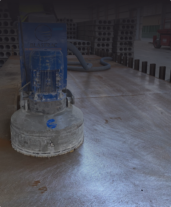 Na podłodze magazynu pracuje maszyna do szlifowania betonowej posadzki. W tle widoczne są ułożone w stosy betonowe rury.