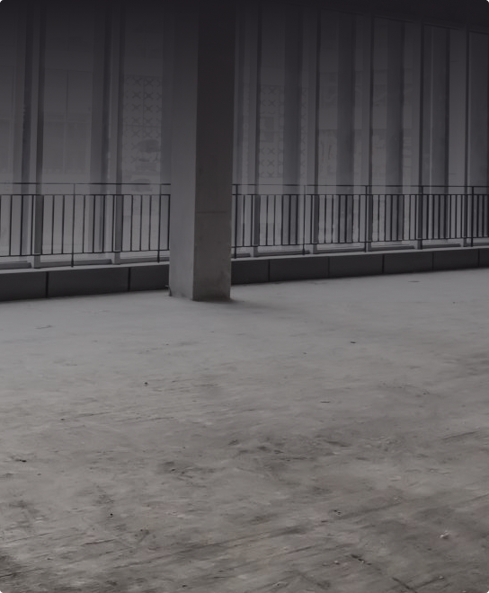 Puste, niedokończone wnętrze z betonową podłogą, pojedynczą kolumną, dużymi oknami i metalową balustradą wzdłuż okien.